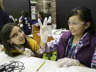 Two young students wearing lab goggles and gloves examine a vial of cloudy water.