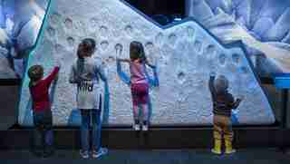Children placing their hands on a display wall of ice bearing many other embedded handprints.